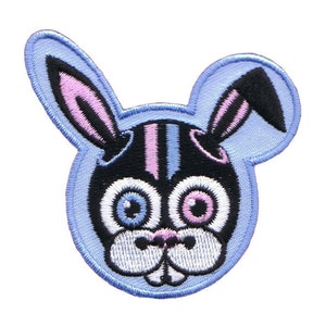 Puede incluir: Un parche bordado azul con la cara de un conejo de dibujos animados. El conejo tiene orejas rosas, pelaje a rayas negro y rosa y grandes ojos azules y rosas.