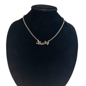 Puede incluir: Un collar plateado con la palabra "Lucky" en cursiva. El collar se exhibe sobre un busto de terciopelo negro, mostrando la delicada cadena y el diseño del colgante. El colgante mide aproximadamente 2,5 cm de largo.