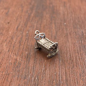May include: A small silver-toned metal charm shaped like a miniature rocking cradle. The charm features a detailed headboard with a crown-like top, a textured side panel, and curved rockers at the base, complete with a small loop for attachment.