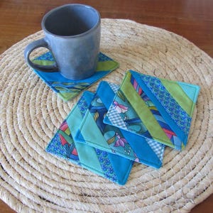 May include: A blue ceramic mug sits on a matching coaster, with several handmade fabric coasters nearby. The coasters have a patchwork design with various blue and green patterned fabrics. The coasters are square and approximately 10 cm by 10 cm.