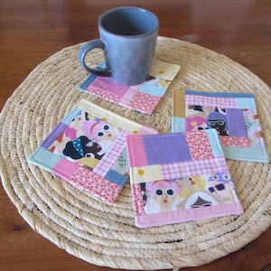 Peut inclure: Ensemble de quatre dessous de verre en tissu faits à la main avec un motif patchwork. Chaque dessous de verre présente un mélange de tissus roses, violets, jaunes et bleus, certains avec un motif répété de visages de dessins animés. Une tasse bleue est posée sur un dessous de verre, le tout sur un dessous de plat tressé.
