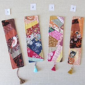 Op de afbeelding: Vier handgemaakte stoffen bladwijzers met kleurrijke patchwork ontwerpen. Elke bladwijzer heeft een ander patroon en kleurenschema, waaronder bloemen-, geometrische en kat-thema prints. Kwasten in verschillende kleuren bungelen aan de onderkant.