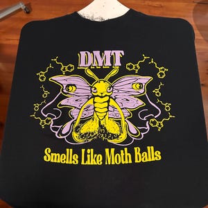 May include: Black t-shirt featuring a yellow and purple moth graphic with the text "DMT" above and "Smells Like Moth Balls" below. The design includes molecular structures. The shirt is displayed on a hanger.