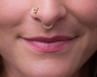 May include: Close-up of a woman's face with a gold nose ring and a small gold stud in her left ear. She is smiling and looking directly at the camera.