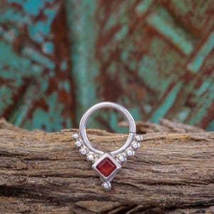 Op de afbeelding: Een zilveren septumring met een rode vierkante edelsteen in het midden. De ring heeft kleine kralenaccenten en een scharnierend ontwerp. De sieraden worden gepresenteerd op een stuk verweerd hout.