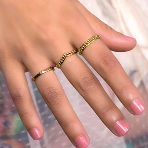 May include: A set of three gold-toned rings. The rings are thin and delicate, with a variety of textures. The rings are worn on the fingers of a hand.