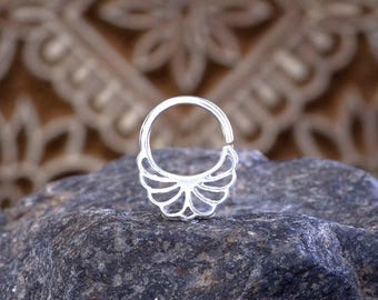 Könnte beinhalten: Ein silberner Septumring mit einem dekorativen, fächerartigen Design. Der Ring ist kreisförmig mit einem Klappverschluss und wird auf einem dunkelgrauen Stein präsentiert. Der Hintergrund ist ein gemusterter Stoff in neutralen Tönen.