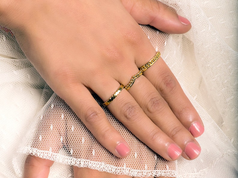 May include: A hand wearing two gold rings. The rings are both thin and have a textured design. The hand is resting on a white lace fabric.