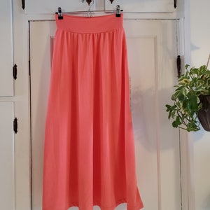 May include: A coral pink midi skirt with a wide waistband and a gathered waistline. The skirt is hanging on a hanger against a white door.