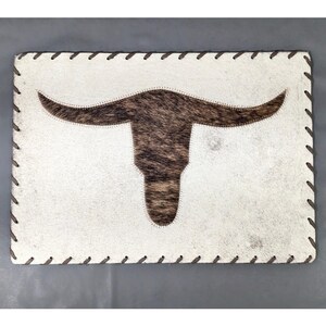 May include: A rectangular placemat featuring a white hair-on-hide background with a centered brown and black mottled cow skull silhouette. The edges are finished with a whip-stitched border using dark brown thread for a decorative finish.