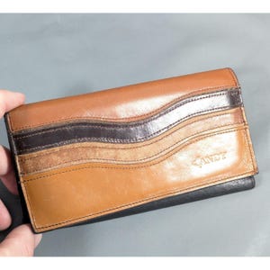 May include: A rectangular leather wallet featuring a multi-toned design with horizontal wavy stripes in shades of tan, medium brown, and dark brown. The word ANDY is embossed in the lower right corner of the front flap, which has a smooth finish.