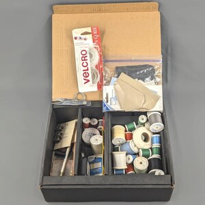 May include: A cardboard sewing supply box containing various spools of thread in assorted colors, a package of white Velcro, a clear plastic bag with beige fabric patches, a small pair of silver metal scissors, and a white marking pencil.