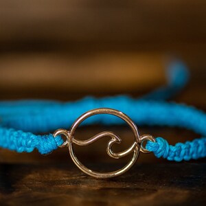 May include: A turquoise macrame bracelet with a gold-colored wave charm. The wave charm is a circular design with a stylized wave in the center. The bracelet is displayed on a dark wooden surface.