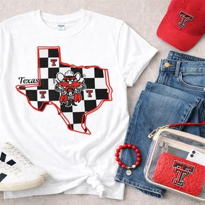 May include: A white t-shirt with a Texas graphic, a red baseball cap, blue jeans, white sneakers, a red bracelet, and a clear crossbody bag with a red insert. The Texas graphic features a black and white checkered design with a red outline.