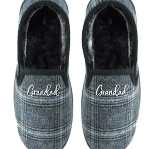 May include: A pair of grey plaid slippers with the word "Grandad" in white script on the top. The slippers have a black trim and a soft, black interior lining. The plaid pattern includes shades of grey and blue.