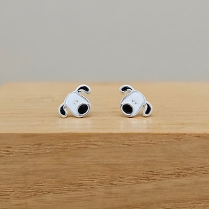 May include: A pair of silver stud earrings featuring a cartoon dog design. The dog is white with black eyes and a black nose.