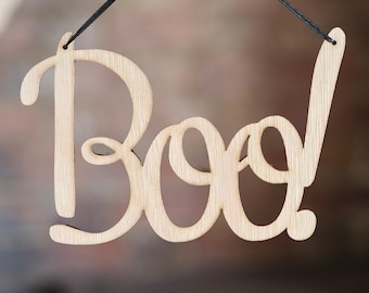 May include: A wooden sign with the word "Boo!" written in a decorative script. The sign is hanging from a black string.