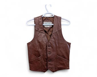 May include: A brown leather vest with a V-neck and three buttons. The vest has two front pockets and is hanging on a white hanger. The label reads "Free Wine".