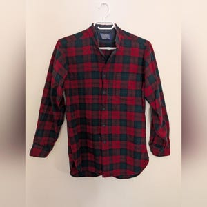 Peut inclure: Une chemise à manches longues à carreaux rouges et verts. La chemise est suspendue à un cintre blanc sur un fond neutre. Le col est de couleur bleu foncé.