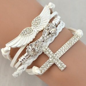 May include: A white multi-strand bracelet featuring silver-tone charms. The charms include a pair of wings, a decorative centerpiece, and a cross, all embellished with clear rhinestones. The bracelet is designed with braided and smooth white cords.