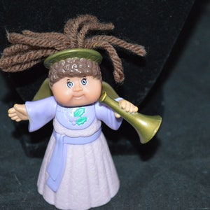 Può includere: Una bambola angelo Cabbage Patch Kid con un vestito viola e un'aureola, che tiene una tromba dorata. La bambola ha i capelli castani e gli occhi azzurri.