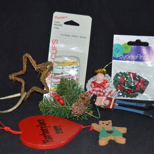 May include: A collection of Christmas ornaments and decorative items. Includes a wooden star, a red heart-shaped ornament with text, a wooden bear, a package of clothespins, and other festive decorations.