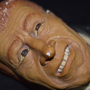 Puede incluir: Un primer plano de una escultura de cer&aacute;mica de un rostro humano. La escultura est&aacute; pintada en tonos marrones y tiene una expresi&oacute;n realista. La boca de la persona est&aacute; ligeramente abierta, revelando un conjunto de dientes.