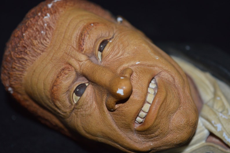 Puede incluir: Un primer plano de una escultura de cer&aacute;mica de un rostro humano. La escultura est&aacute; pintada en tonos marrones y tiene una expresi&oacute;n realista. La boca de la persona est&aacute; ligeramente abierta, revelando un conjunto de dientes.