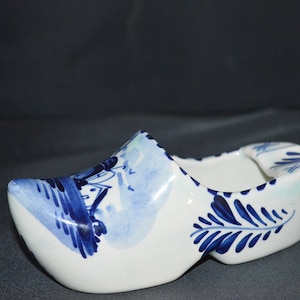 May include: A white ceramic clog-shaped ashtray with blue floral and landscape designs. The ashtray features a detailed painted design on the front and sides, with a smooth, glossy finish. The interior is open for use.