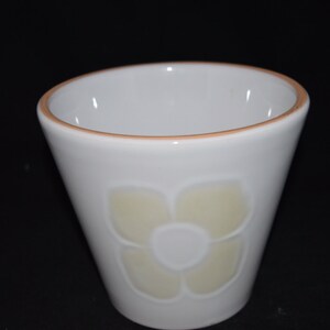 May include: A white ceramic cup with a light yellow flower design. The cup has a brown rim.