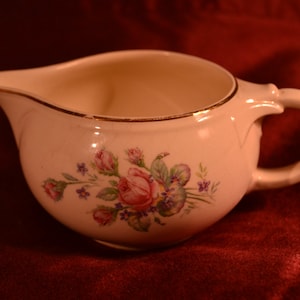 Peut inclure: Un pot à crème en céramique blanche avec un bord doré et un motif floral composé de roses roses et de fleurs violettes.