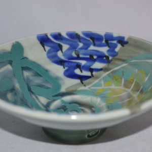 May include: A small, white ceramic bowl with a footed base. The bowl is decorated with abstract designs in shades of blue, green, and yellow.