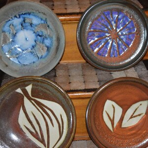 May include: Four ceramic plates with different designs. The first plate has a blue and white abstract design. The second plate has a purple and brown flower design. The third plate has a white and brown abstract design. The fourth plate has a brown and white leaf design.