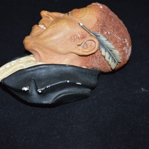 Puede incluir: Un busto de cer&aacute;mica de un hombre con una pluma en el pelo. El busto est&aacute; pintado en tonos marrones, negros y blancos. El hombre sonr&iacute;e y tiene una pluma en el pelo.