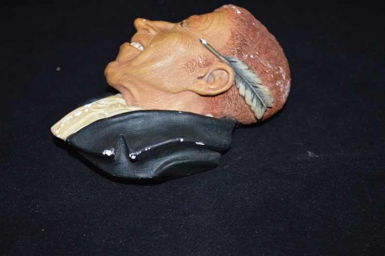 Puede incluir: Un busto de cer&aacute;mica de un hombre con una pluma en el pelo. El busto est&aacute; pintado en tonos marrones, negros y blancos. El hombre sonr&iacute;e y tiene una pluma en el pelo.