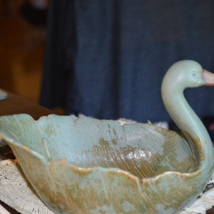 May include: A ceramic swan-shaped bowl with a green glaze. The bowl has a leaf-like design and is sitting on a white cloth.