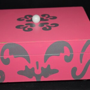Puede incluir: Una caja de madera rosa con un diseño floral gris y una perilla blanca en la tapa.