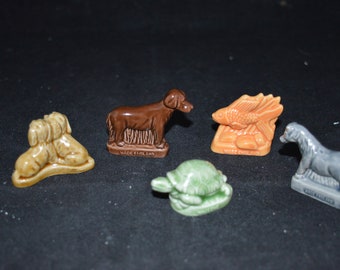 Può includere: Una collezione di piccole statuette di animali in ceramica, tra cui un cane marrone, un cavallo grigio, una tartaruga verde, una coppia di cani beige e un pesce arancione. Ogni statuetta è dettagliata e poggia su una piccola base. Le parole "WADE ENGLAND" sono visibili su alcune basi.