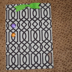 May include: A beige cork board with a black and white geometric pattern. The board has a green bow at the top and three colorful push pins.