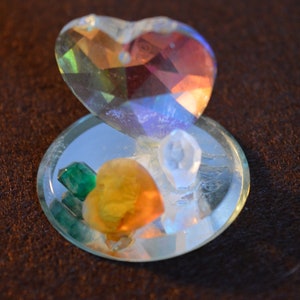 以下が含まれることがあります： 鏡面台座が付いたハート型のクリスタルオーナメント。台座には、緑、オレンジ、白など、さまざまな小さなクリスタルが入っています。