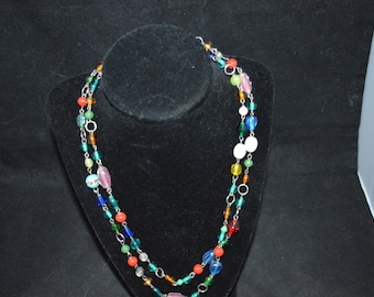 May include: A colorful beaded necklace with a variety of shapes and sizes. The necklace features beads in shades of blue, green, orange, pink, and white, strung on a silver chain. The necklace is displayed on a black velvet bust.