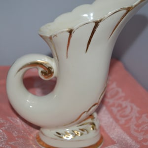 Może przedstawiać: Biały ceramiczny wazon z pozłacanym wykończeniem w kształcie rogu. Wazon ma zakrzywiony uchwyt i ozdobną podstawę.