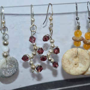 May include: Three pairs of earrings with different styles. The first pair features white beads and silver glitter. The second pair has purple crystal beads and silver wire. The third pair has brown and orange beads with a large, round, white button.