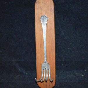 May include: A wooden wall-mounted coat rack with a silver fork attached as a hook. The fork has a decorative design at the top.