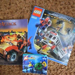 May include: Three LEGO instruction manuals for building sets. The first manual is for LEGO Dino Attack set 7475, featuring a red and black off-road vehicle with a dinosaur. The second manual is for LEGO Spider-Man 2 set 4855, featuring Spider-Man and Doc Ock. The third manual is for LEGO System set 6110, featuring a green and black underwater creature.