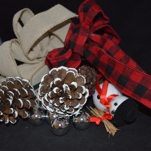 Könnte beinhalten: Eine Sammlung von Weihnachtsdekorationen, darunter ein rot-schwarzes Karoribbon, eine braune und weiße Tannenzapfen, eine kleine Schneemannfigur und silberne Ornamente.