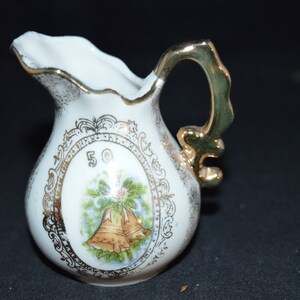 Puede incluir: Una jarra de porcelana blanca con asa y ribetes dorados. La jarra tiene un diseño dorado con dos campanas y el número 50.