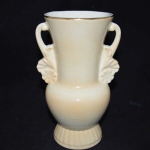 May include: A cream-colored ceramic vase with gold trim and leaf-shaped handles. The vase has a rounded body and a flared base.