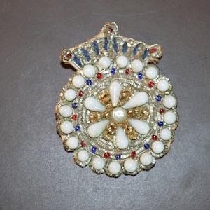 May include: A white and gold beaded brooch with a crown design and a central pearl. The brooch is decorated with red and blue beads.