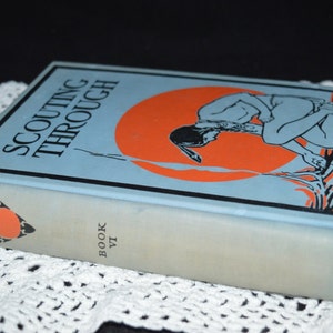 Può includere: Libro vintage intitolato "Scouting Through" con copertina azzurra e un'illustrazione di una figura nativa americana contro un cerchio arancione. Il libro è appoggiato su un centrino di pizzo bianco.